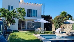 Una casa moderna de dos pisos con piscina, un coche aparcado en la entrada y una zona de patio con mobiliario exterior.
