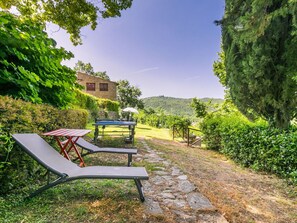 A stone pathway leading to a table and chairs in a lush garden setting, with a house in the background.
