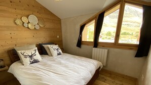 Un dormitorio con techo inclinado de madera, una cama con ropa de cama blanca, dos almohadas con estampado de ciervo, una mesita de noche de madera y una ventana con vistas a un paisaje montañoso.