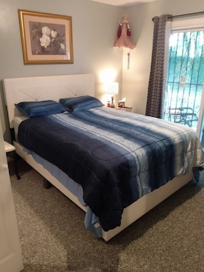 A bedroom with a bed, a nightstand, a lamp, a framed picture, and a window with curtains.