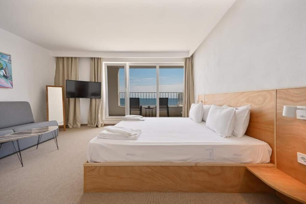 Habitación de hotel con cama, televisión, balcón con vistas y zona de estar con mesa y sillas.