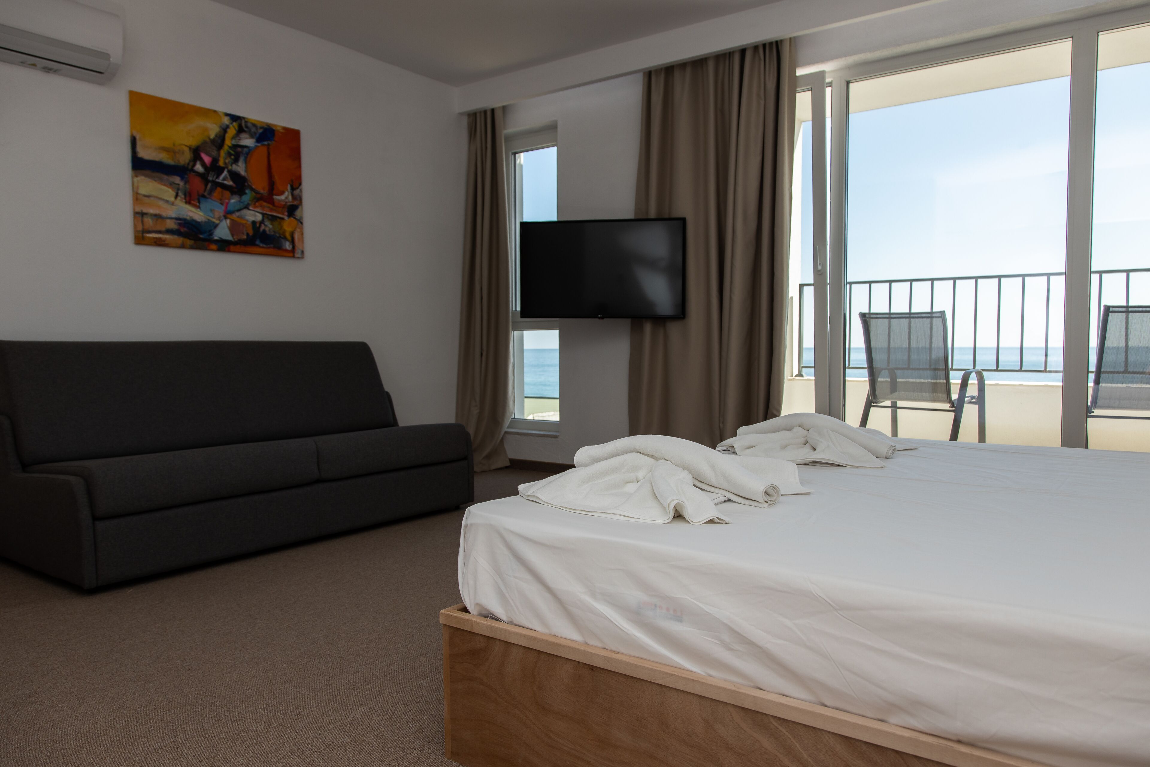 Quarto de hotel com cama, sofá, televisão e varanda com vista para o mar.