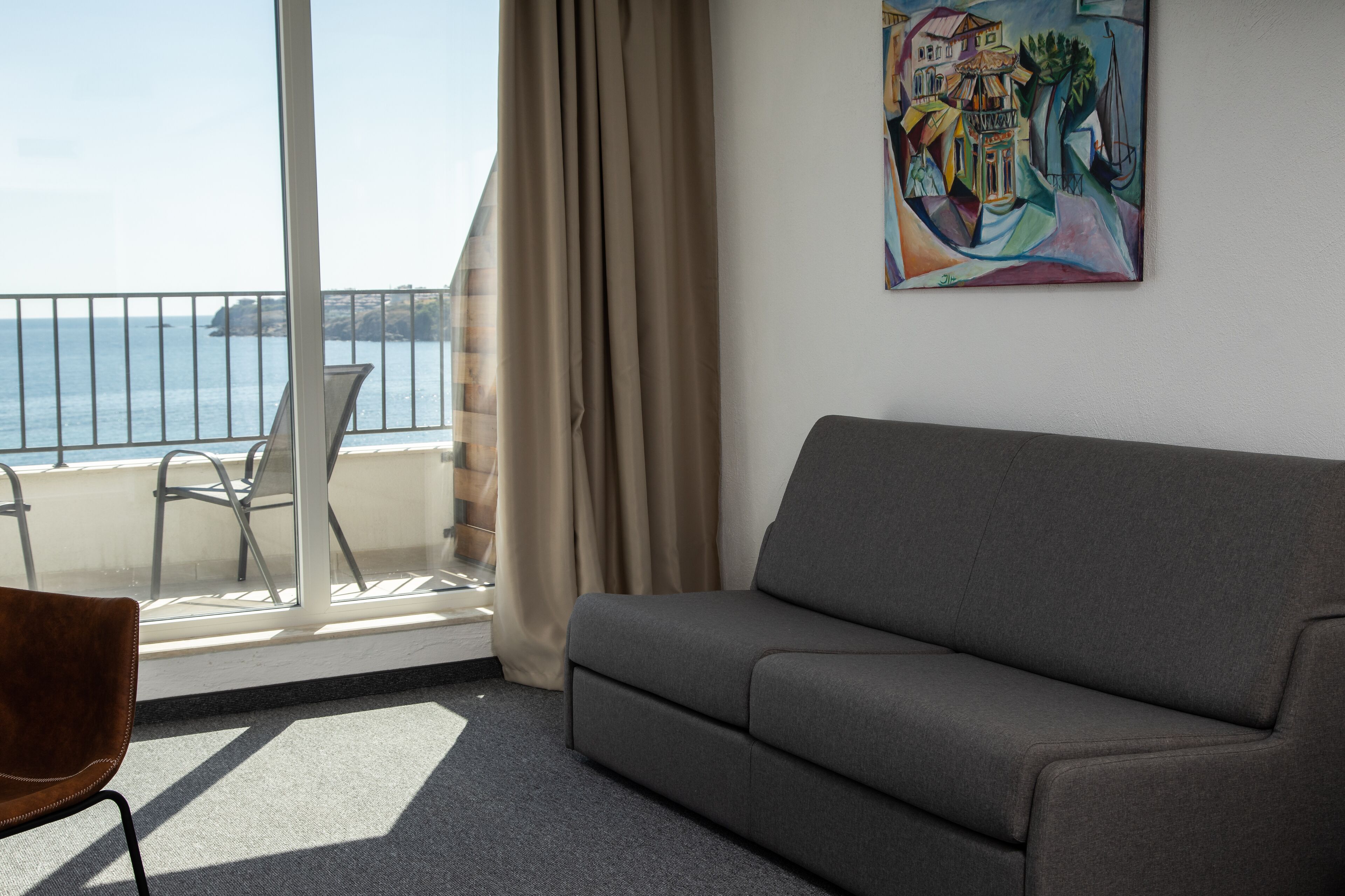 A room with a grey sofa, a chair, a painting, a window with a view of the sea, and a curtain.