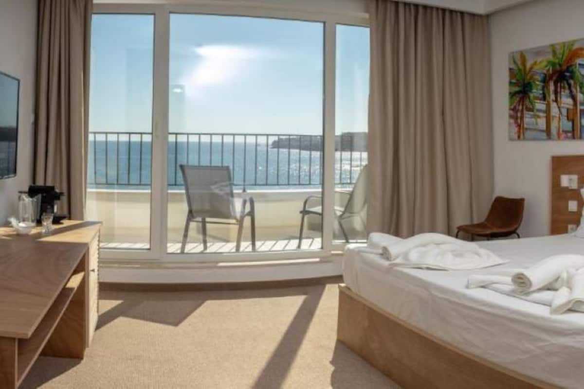Habitación de hotel con cama, escritorio, televisor, balcón con silla y un cuadro.