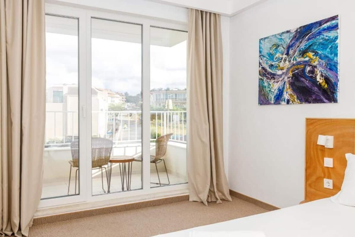 Una habitación de hotel con un gran ventanal corredero que da a un balcón, una cama con cabecera de madera y una obra de arte abstracta en la pared.
