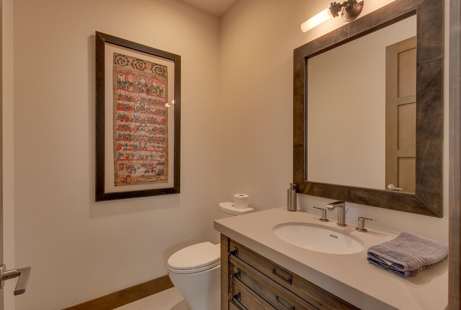 A bathroom with a large framed picture, a sink, a mirror, and a toilet.