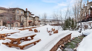 A snow-covered outdoor area with picnic tables and benches, a stone wall, and a building with multiple windows and a brick facade. - Guest-Favorite Ski Home in Bear Hollow w/ Hot Tub – Sleeps 18 – King/Queen Beds (Park City)