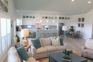 A modern living room with a beige sofa, a coffee table, and a patterned armchair. A kitchen with white cabinets and a dining area with a table and chairs is visible in the background. - Pelican Wharf, Pool/Hot Tub (Crystal Beach)