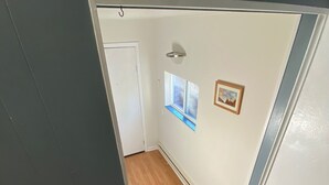 Un couloir étroit au plancher de bois, une porte blanche, une petite fenêtre donnant sur le paysage et un tableau accroché au mur.