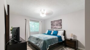 A bedroom with a bed, a desk with a computer, a window with curtains, a ceiling light, and a painting on the wall.
