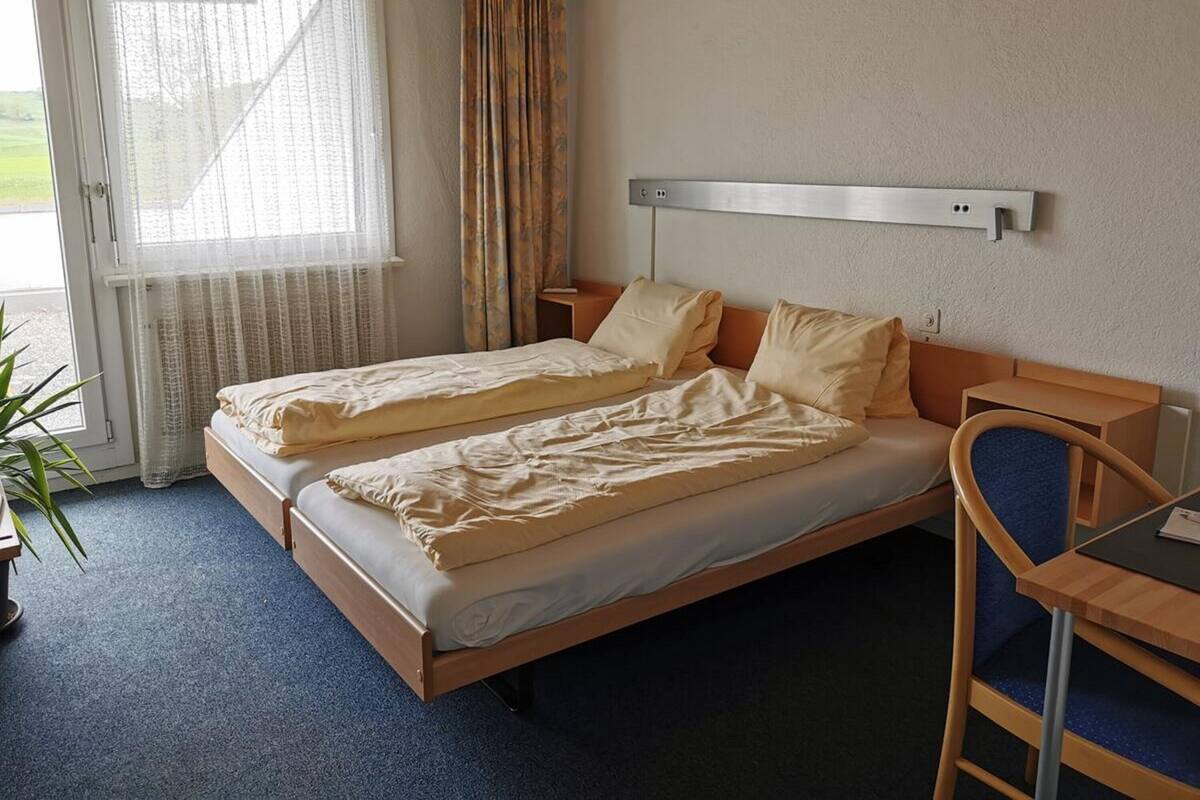 Una cama doble con estructura de madera, una mesita de noche, una silla y una ventana con cortinas.