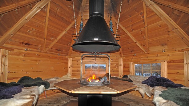 L’intérieur d’une cabane en rondins, avec un poêle à bois central, des murs en bois et une fenêtre donnant sur l’extérieur.