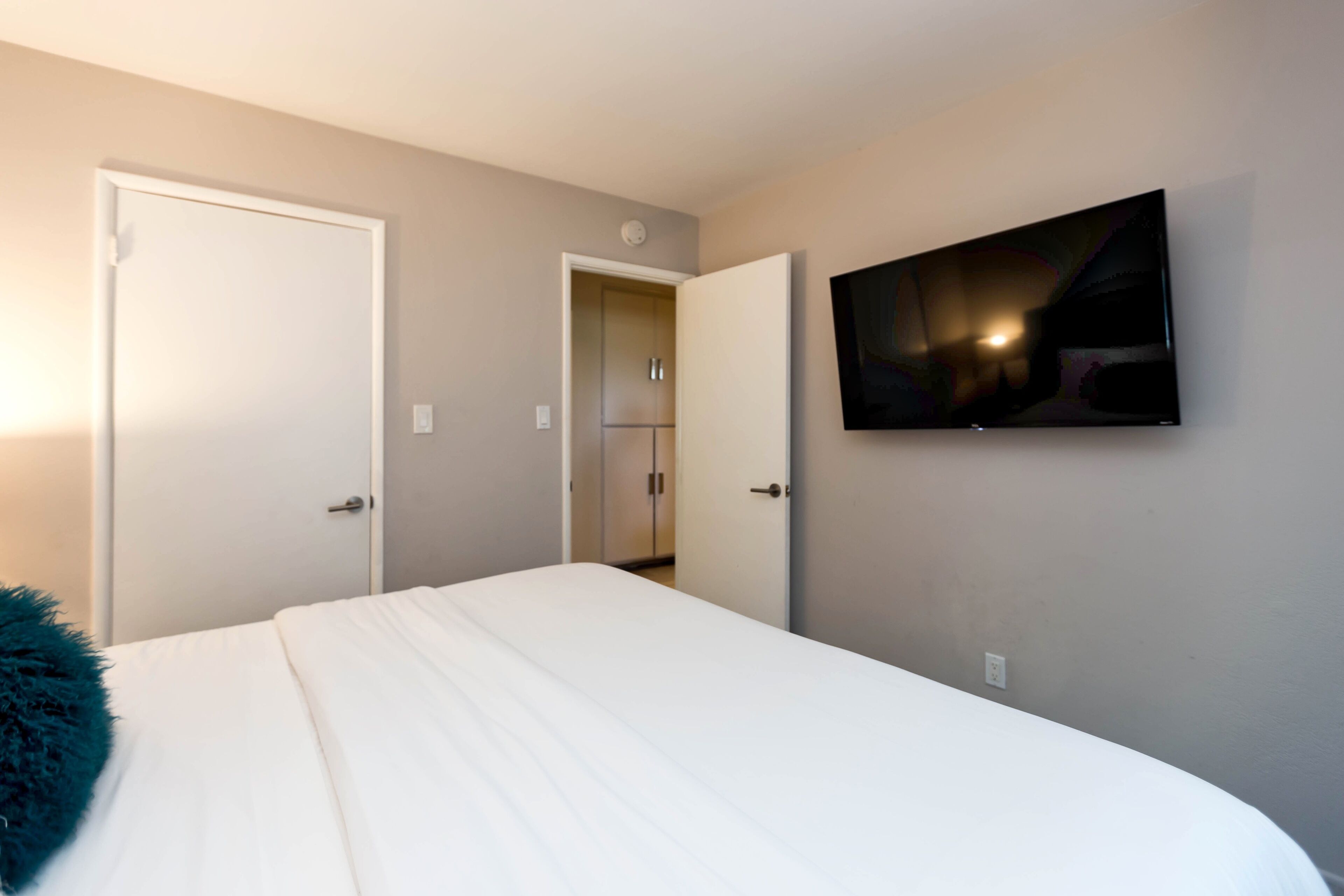 Ein Schlafzimmer mit einem Bett, einem an der Wand befestigten Fernseher und zwei Türen zu anderen Räumen.