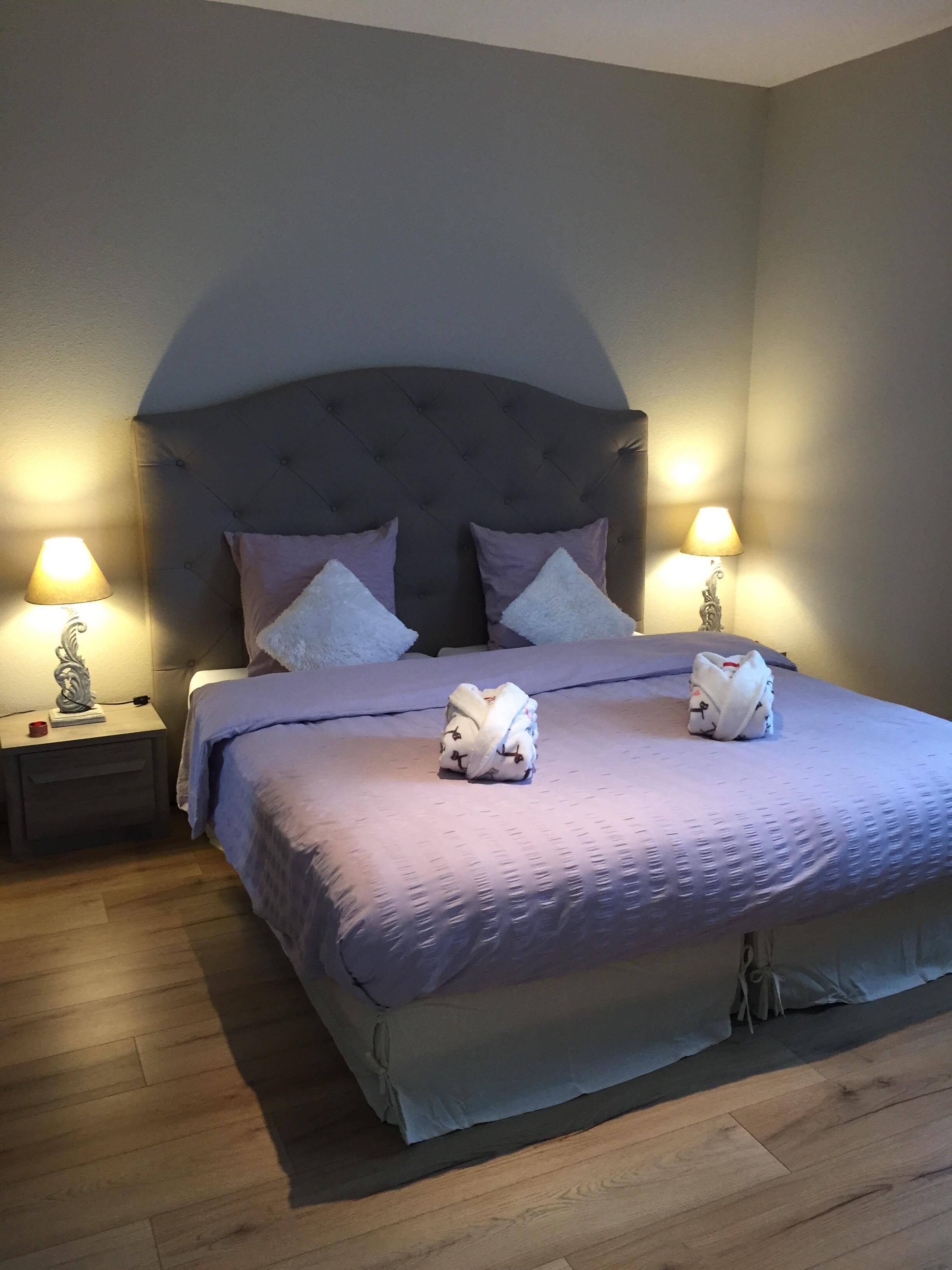 A neatly made bed with a tufted headboard, two pillows, and two folded towels. There are two bedside tables with lamps and a small decorative item on one. The room has wooden flooring and light-colored walls.