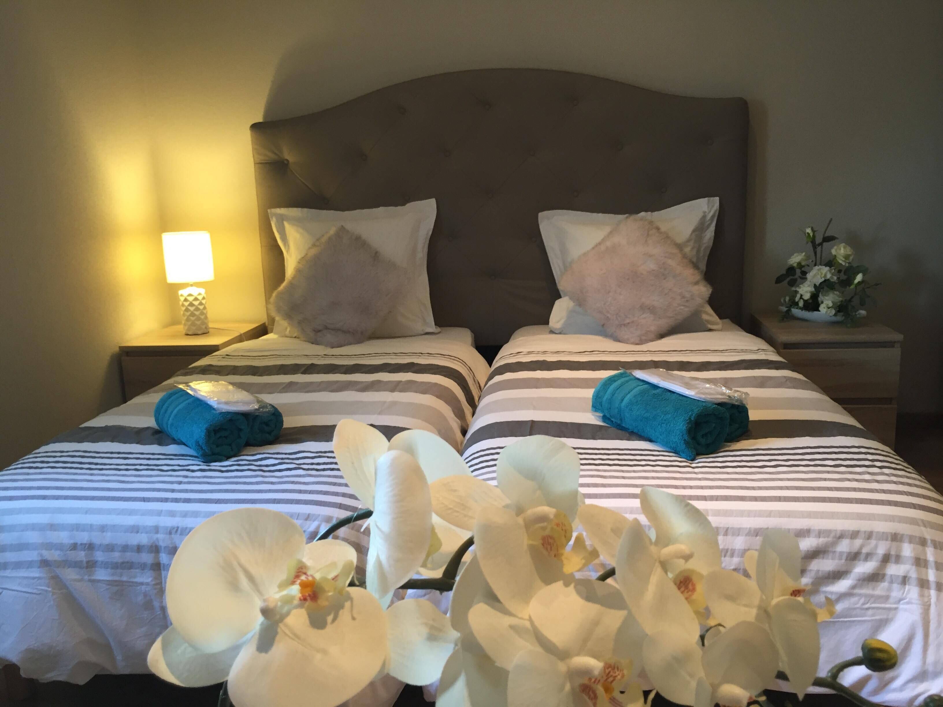 Habitación de hotel con dos camas, ropa de cama a rayas, cabecero, mesita de noche con lámpara y una pequeña mesa con flores.