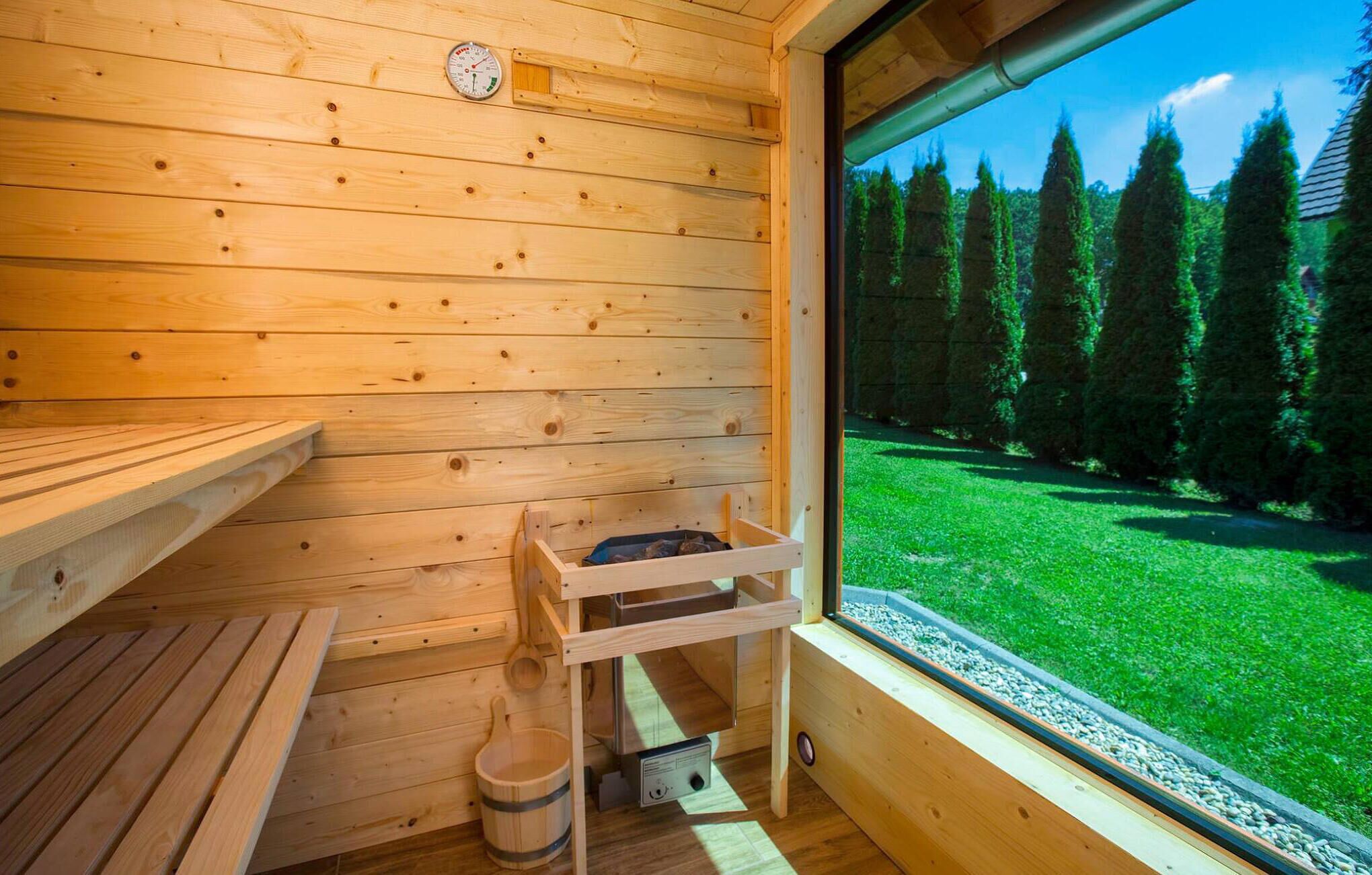 A wooden sauna room with a bench, a wooden bucket, and a window overlooking a forest.
