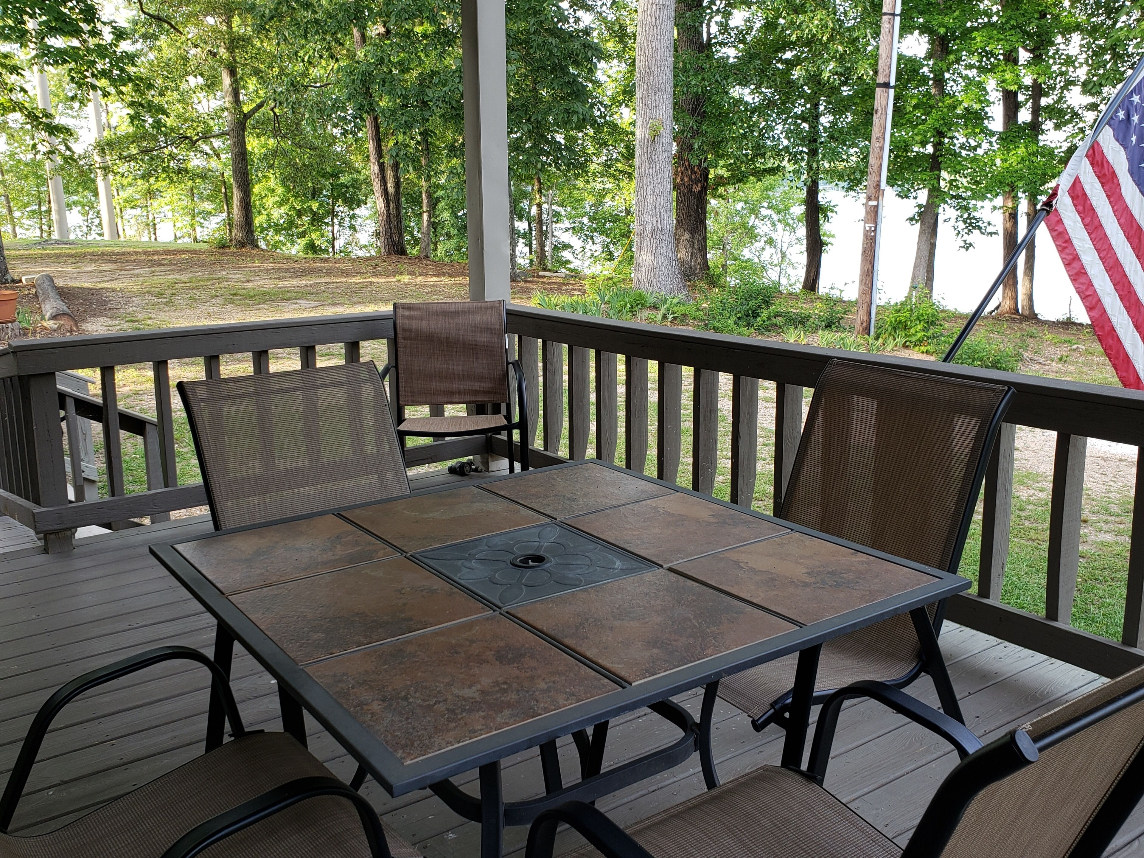Une terrasse extérieure couverte, avec une table et des chaises, entourée d’une terrasse en bois et offrant une vue sur les arbres.