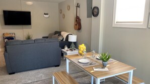 A modern living room with a grey sofa, a wooden dining table with benches, a TV mounted on the wall, a guitar, and a clock. - Private Nashville Guest Suite - King Bed - Massage Chair - Private Entry (Hermitage)