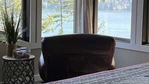 Een slaapkamer met een groot bed, een stoel, een nachtkastje en uitzicht op een meer en bomen door de ramen.