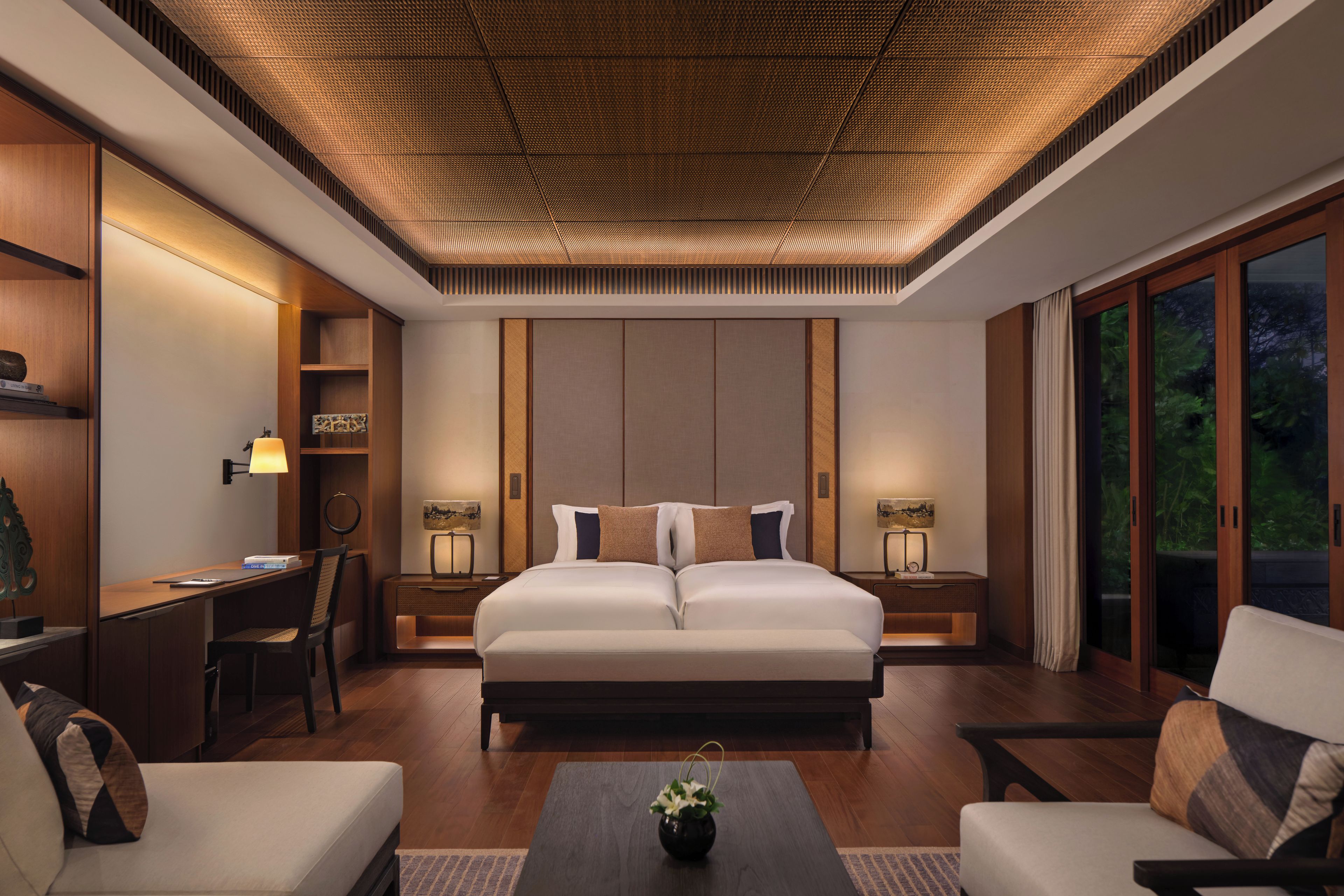 A modern bedroom with a large bed, bedside tables, a desk, and a view of greenery through the windows.