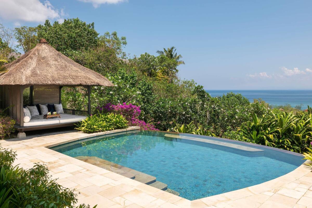 A swimming pool with a thatched-roof gazebo, surrounded by lush greenery and a view of the ocean.