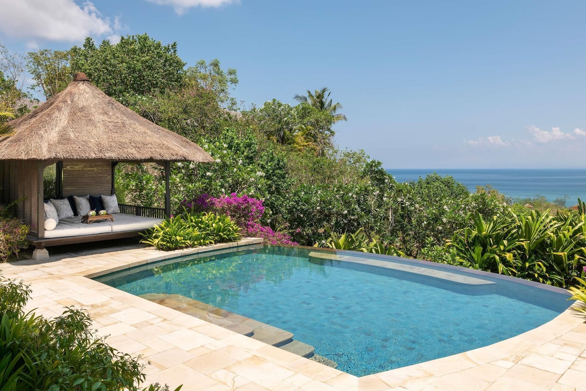 A swimming pool with a thatched-roof gazebo, surrounded by lush greenery and a view of the ocean.