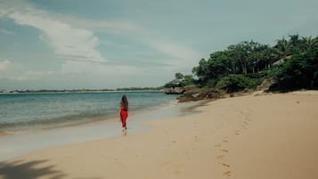 Una playa con una persona caminando sobre la arena, cielo despejado y mar tranquilo.