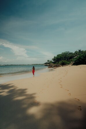 A beach with a person walking on the sand, a clear sky, and a calm sea. - Raffles Bali (Jimbaran)