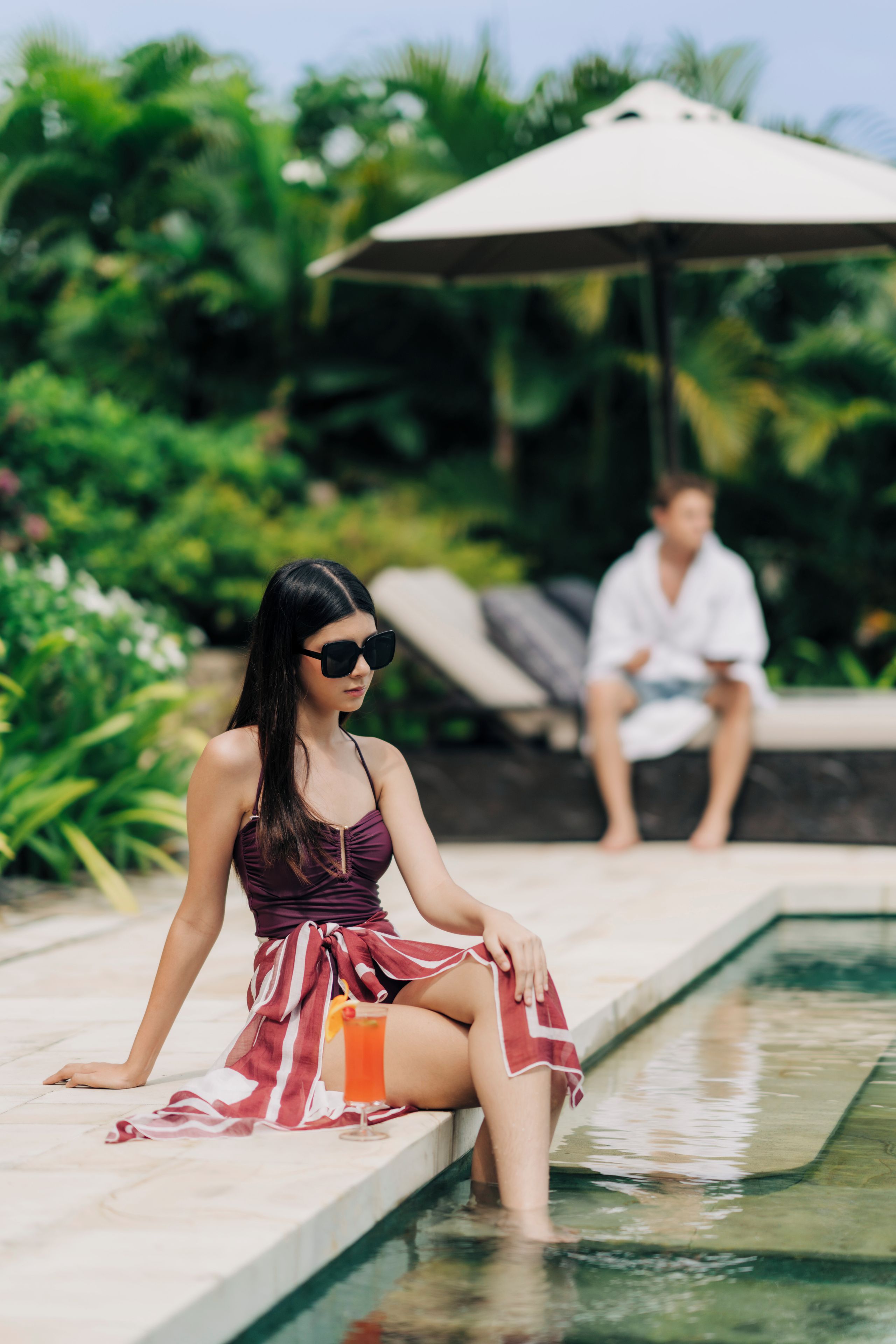 A woman sitting by a pool with a man in the background.
