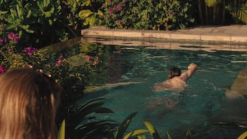 Una persona sentada junto a la piscina leyendo un libro, rodeada de exuberante vegetación y una piscina cristalina.