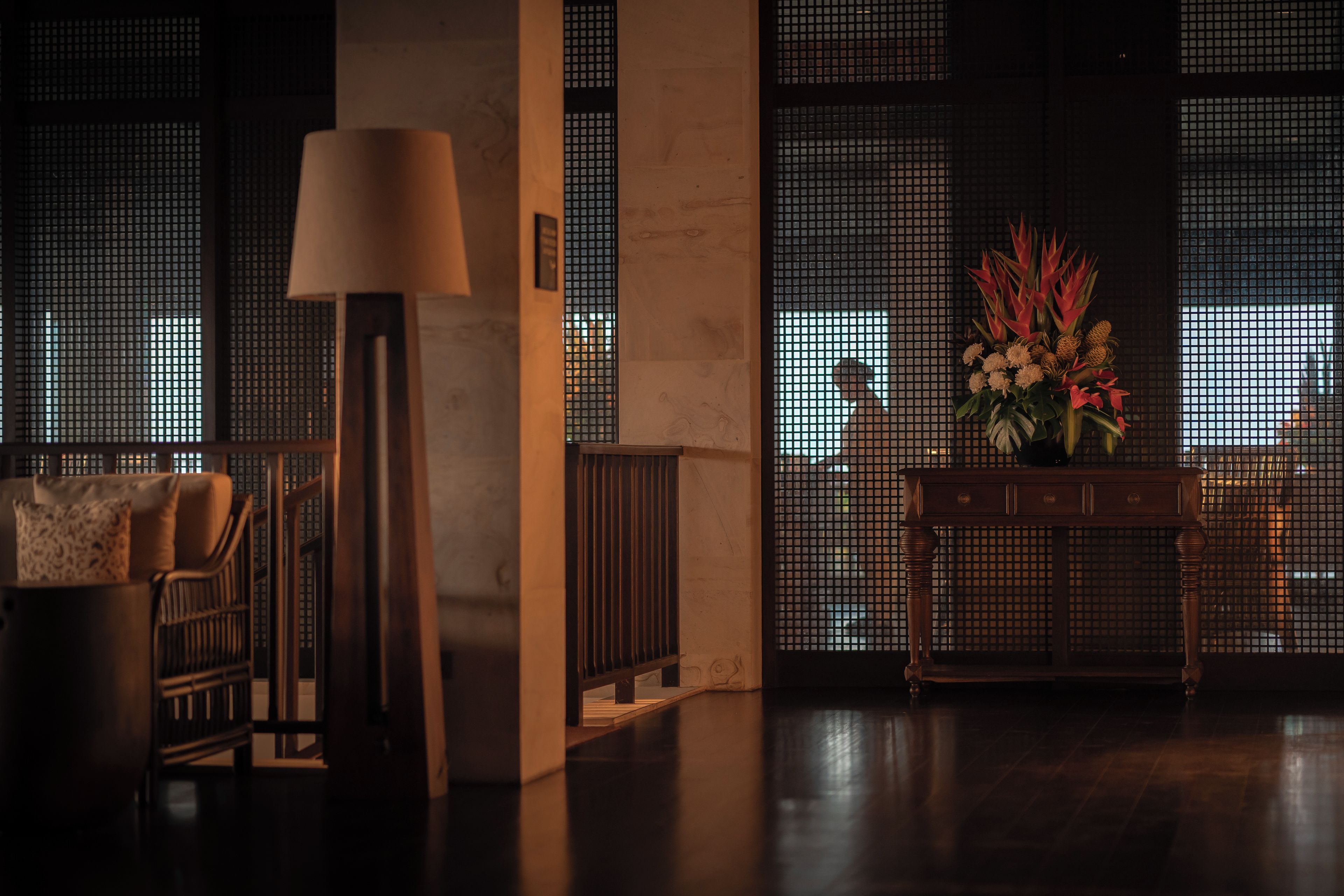 Una habitación con poca luz, un sofá, una lámpara, una consola con un arreglo floral y vistas a través de una ventana con diseño de cuadrícula.