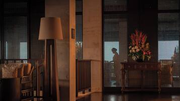 Una habitación con poca luz, un sofá, una lámpara, una consola con un arreglo floral y vistas a través de una ventana con diseño de cuadrícula.