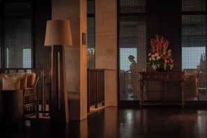 A dimly lit room with a sofa, a lamp, a console table with a floral arrangement, and a view through a window with a grid pattern. - Raffles Bali (Jimbaran)