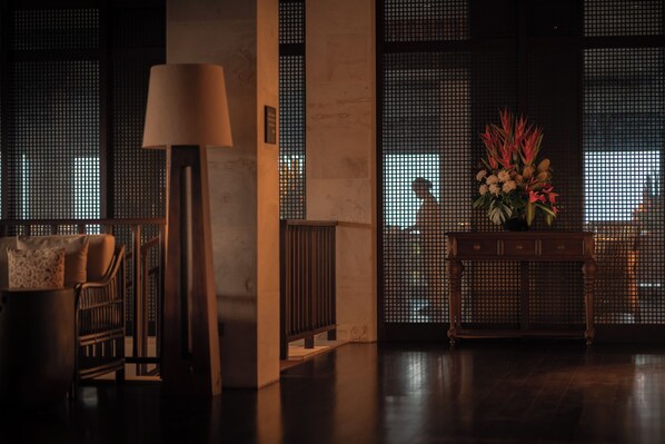 A dimly lit room with a sofa, a lamp, a console table with a floral arrangement, and a view through a window with a grid pattern.