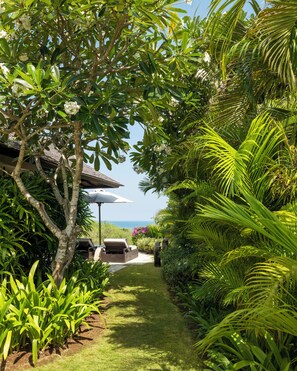 A garden pathway leading to a building with a thatched roof, surrounded by lush greenery and a clear sky. - Raffles Bali (Jimbaran)