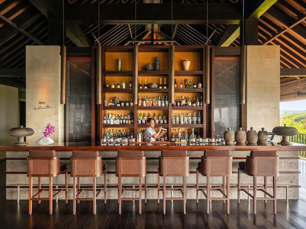 A bar area with multiple shelves of bottles, a bartender, and wicker bar stools. - Raffles Bali (Jimbaran)