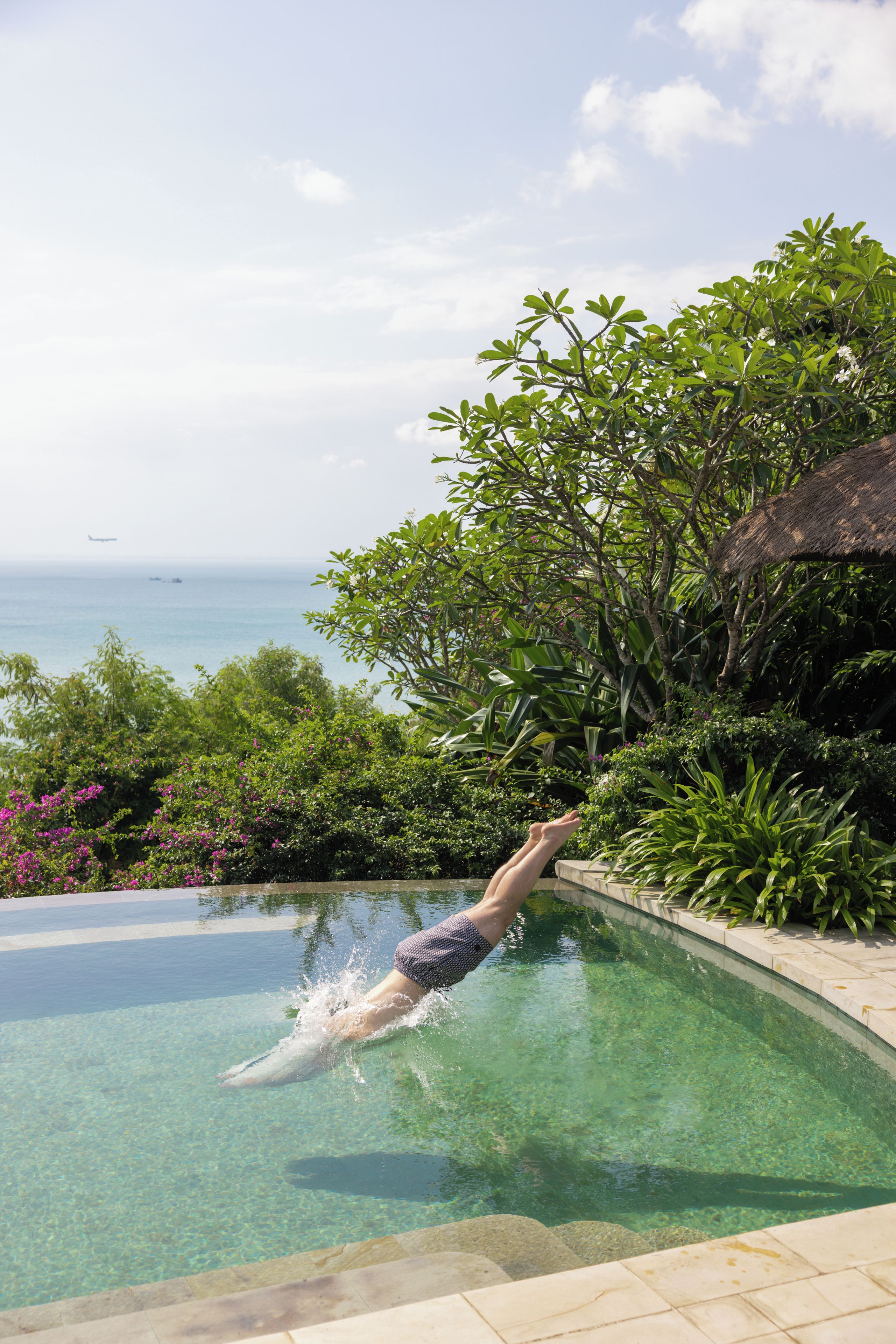Una persona se sumerge en una piscina cristalina con vistas al océano y exuberante vegetación.