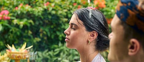 A person pouring water over another person's head, with greenery in the background.