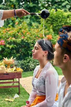 A person pouring water over another person's head, with greenery in the background. - Raffles Bali (Jimbaran)