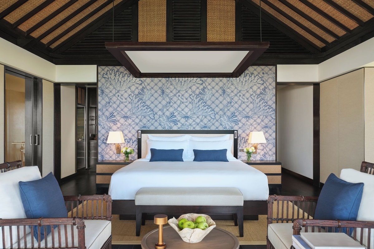 Habitación de hotel con una cama grande, dos mesitas de noche con lámparas, una mesa redonda con frutero y un papel tapiz estampado.