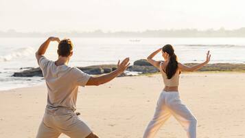 Dos personas practican Tai Chi en una playa arenosa, con el océano al fondo.