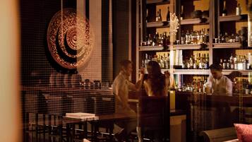 A bar area with a patterned wall decoration, shelves of bottles, and a seating arrangement with chairs and tables.