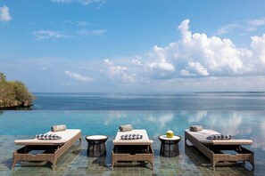 A poolside lounge area with four sunbeds, a small round table, and a lounge chair. - Raffles Bali (Jimbaran)