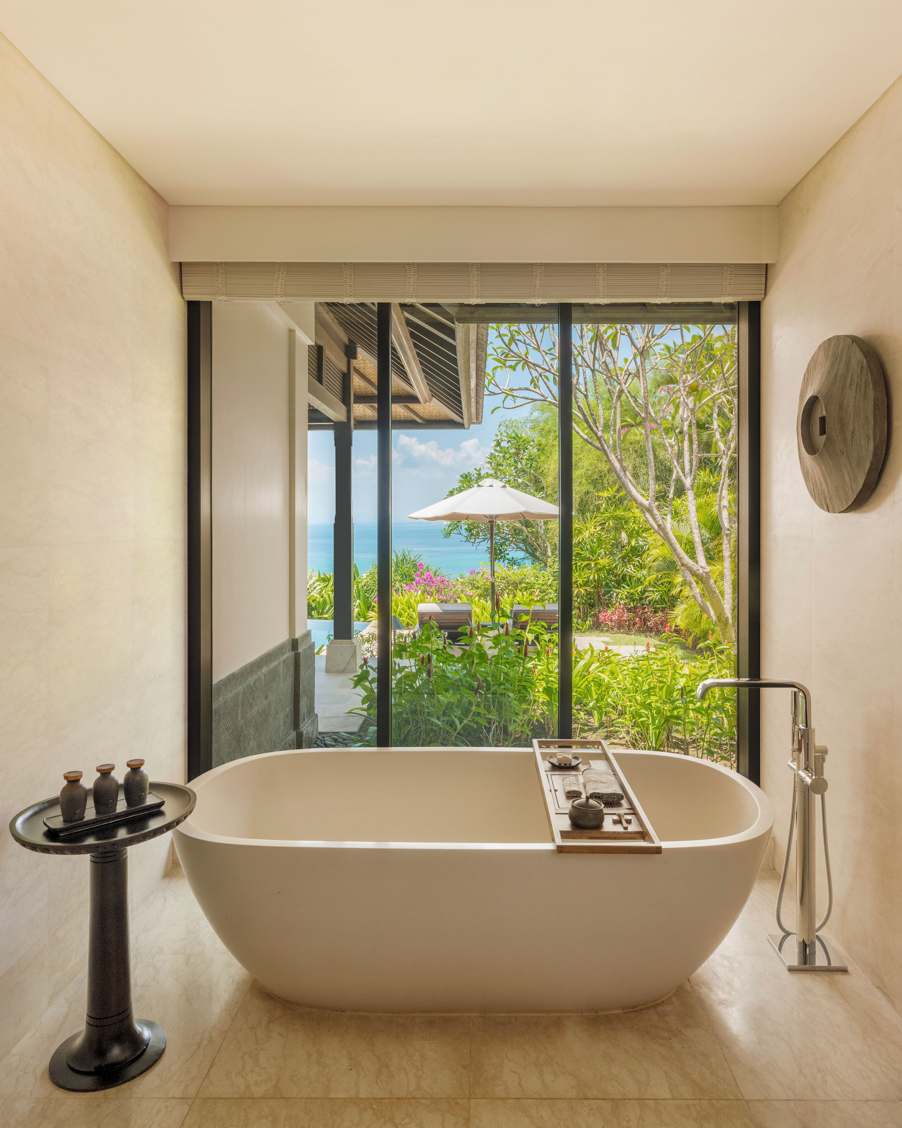 A modern bathroom with a freestanding bathtub, a small table with bottles, and a view of a garden and ocean through large windows.
