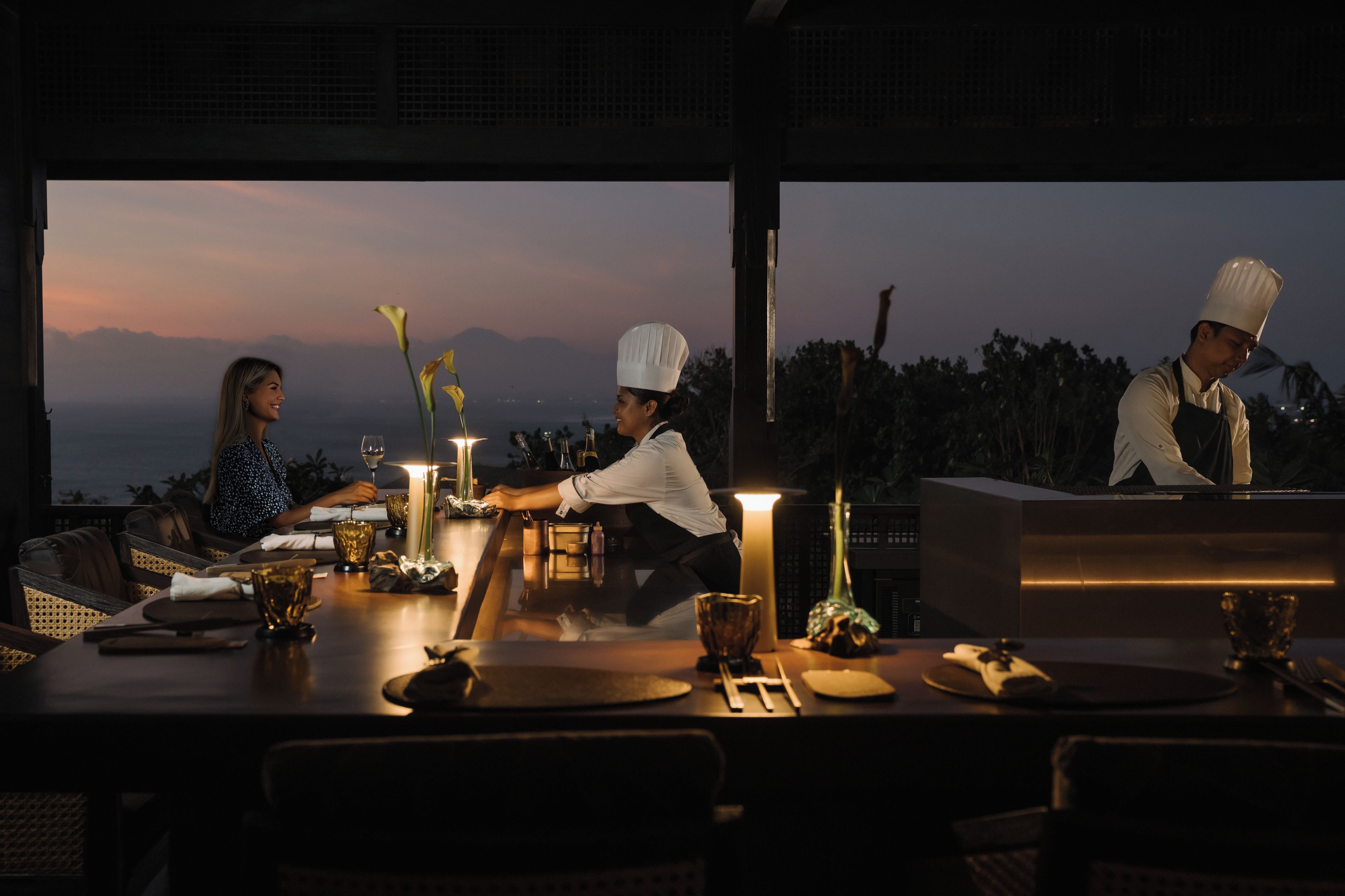 A dining table set for two with a candle, a vase with flowers, and a view of the mountains at dusk.