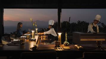 A dining table set for two with a candle, a vase with flowers, and a view of the mountains at dusk.