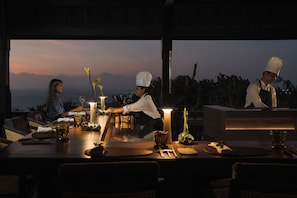 A dining table set for two with a candle, a vase with flowers, and a view of the mountains at dusk.