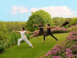 Three people practicing yoga in a grassy outdoor area with trees and flowers.