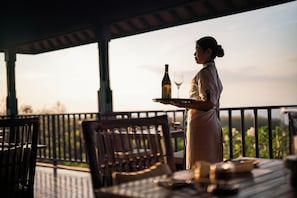 A person carrying a tray with a bottle and a glass, standing on a balcony with a table and chairs, overlooking a landscape. - Raffles Bali (Jimbaran)