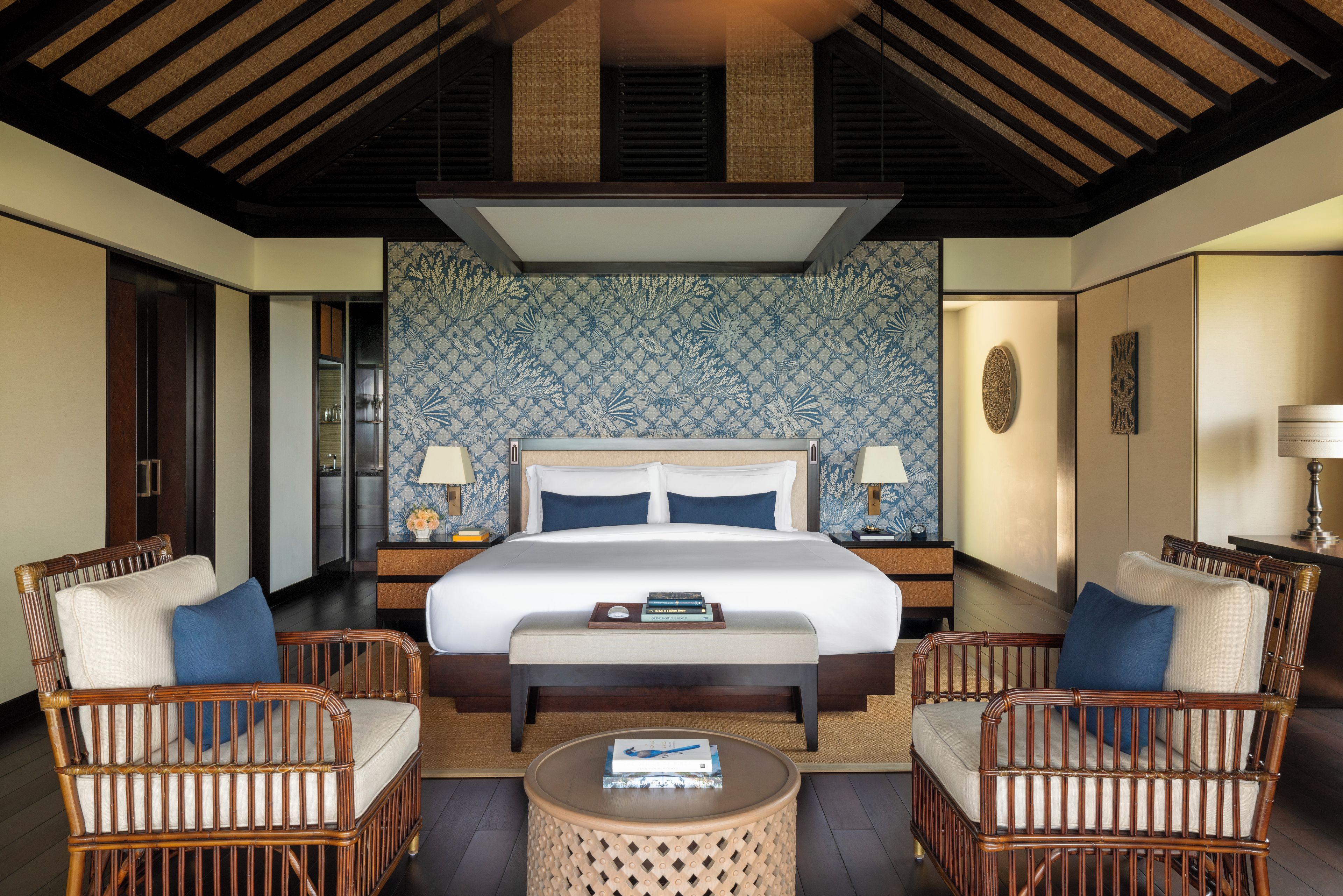 A hotel room with a large bed, two bedside tables, two lamps, a round coffee table, and two wicker chairs.